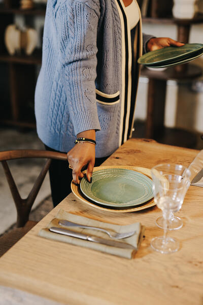 A person in a blue sweater sets green plates on a wooden dining table with cutlery and glassware, preparing for a meal in a cozy, warmly-lit room.