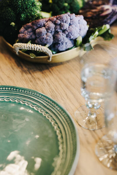 A decorative green plate sits on a wooden table beside a blurry glass, with a basket containing purple cauliflower and green broccoli in the background.