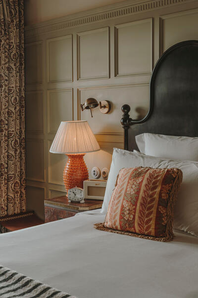 A neatly made bed with a patterned throw pillow and white sheets beside a bedside table, holding a textured lamp, vintage radio, and an alarm clock, all in a well-decorated room.