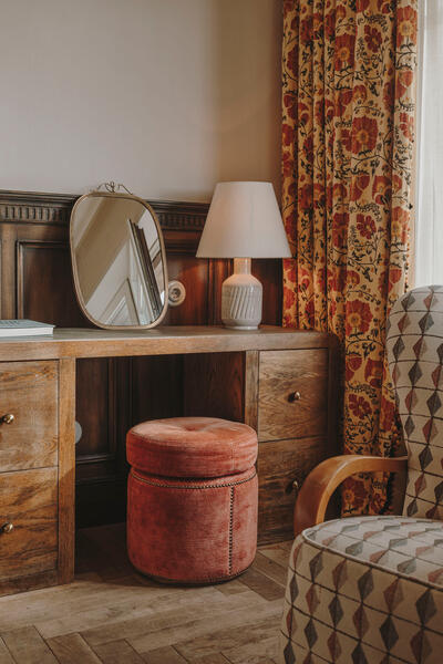 A wooden desk holds a mirror and lamp; a red cushioned stool sits beneath. The setting is a cozy room with floral curtains and a patterned armchair.