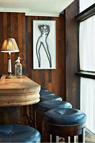 Framed abstract artwork of a human figure hangs on a wooden-paneled wall inside a cozy bar with a raw wooden counter, black cushioned stools, a lamp, and a seltzer water bottle.