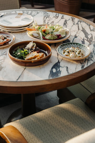 Plates of assorted food, including vegetables and cheeses, rest on a round, marble-topped table with wooden edges, with cushioned dining chairs positioned around it.