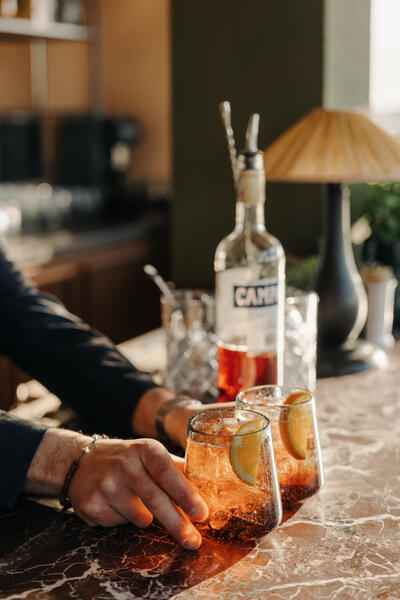 Two glasses with an orange-hued drink and lemon slices are being placed on a marble countertop by a hand; the background shows a bottle labeled "Campari," glassware, and a table lamp.