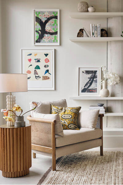 A beige armchair with a patterned cushion and round side table sits in a modern living room with bookshelves, artwork, and minimalist decor on a textured rug.