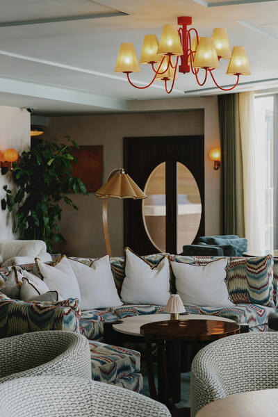 A patterned sofa set with white pillows is placed around a dark wooden table, under a red chandelier, in a cozy, well-lit living room with large oval windows.