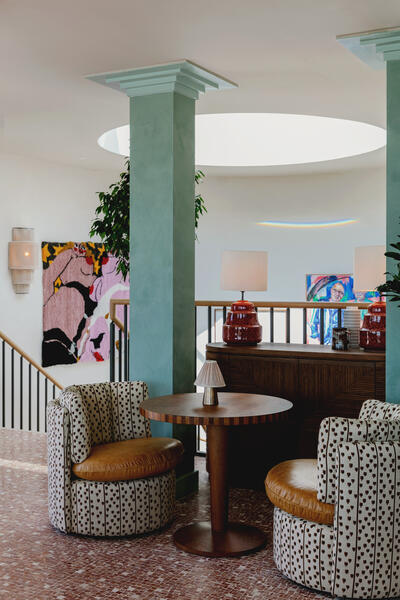 Two patterned armchairs and a wooden table in a modern, open interior with teal columns, art on the walls, red lamps on a console, and a large circular skylight.