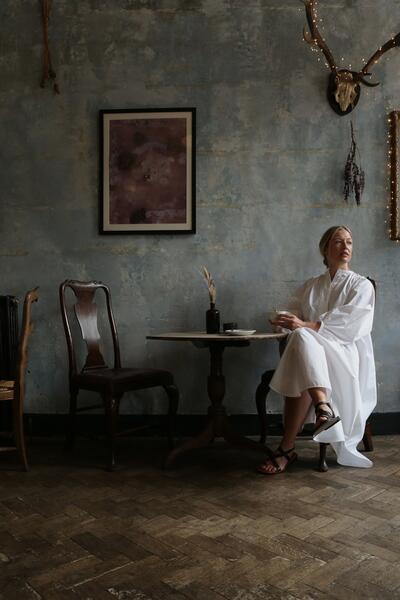 A woman in a white dress sits at a small wooden table in a rustic room with a framed picture, dried plants, and a deer skull with lights on the wall.