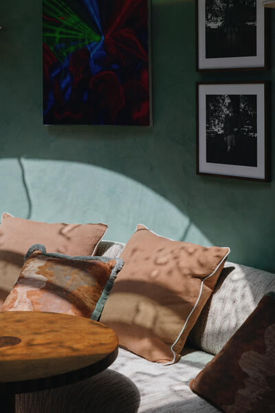 Tan pillows rest on a light grey couch beside a wooden coffee table, in a room with green walls decorated with a colorful abstract painting and two black-and-white photographs. Sunlight filters in, casting shadows.