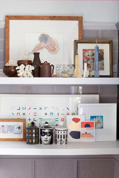 Art-filled shelves showcase framed drawings, ceramic vessels, two model skyscrapers, and decorative jars beside geometric and illustrative prints, all arranged against a tiled wall in a calm domestic setting.