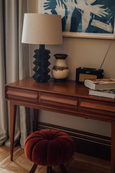 A wooden table displays a modern black lamp, a ceramic vase, and a radio beside stacked books in a softly lit room with a framed abstract hand artwork and beige curtains.