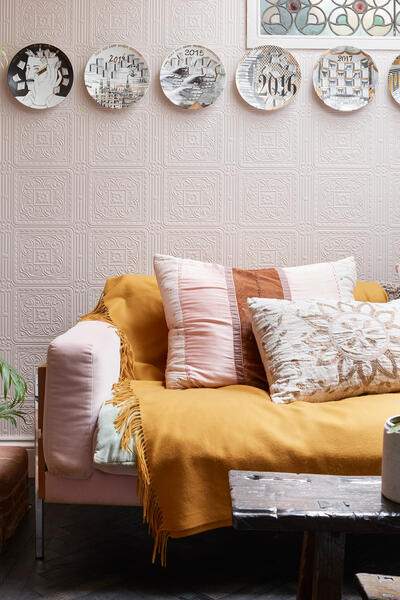 A cozy living room features a pink sofa adorned with cushions and a mustard throw blanket, set against a patterned wall with decorative plates. Indoor plants and a brick fireplace complete the scene.