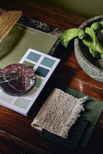 Assorted fabric swatches, color samples, a round stone, and green foliage sit on a wooden table. Text reads "Lick x SOHO HOUSE." Various shades of green color samples labeled "GREEN 03," "GREEN 04," and "GREEN 05."