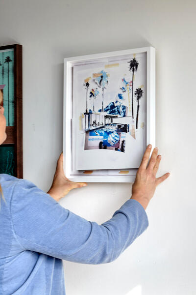 A person with long blonde hair adjusts a framed artwork on a white wall, surrounded by two other framed pictures.