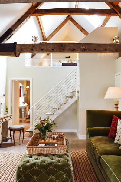 A cozy living room with green velvet furniture, a wicker basket on an ottoman, and a staircase leading to a loft area with exposed wooden beams and soft lighting.