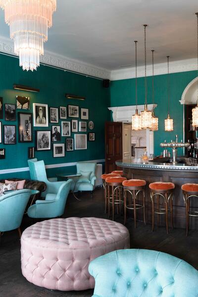 A cozy bar features teal walls adorned with framed pictures, an assortment of light blue and pink tufted chairs, and a central bar surrounded by orange stools.