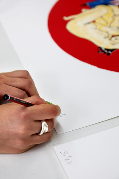 A hand wearing a ring writes "5/25" and another word on white paper using a pencil. Nearby is an illustration featuring a red circular background with a colorful drawing.