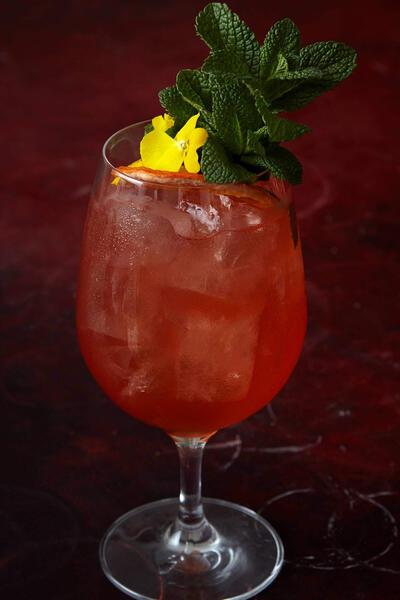 A stemmed glass holds an iced red beverage garnished with a mint sprig and a yellow flower, set against a dark red, textured background.