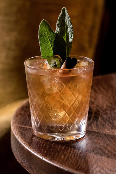 A cocktail with ice cubes and garnished with mint leaves sits in a diamond-patterned glass on a wooden table beside a golden cushion.