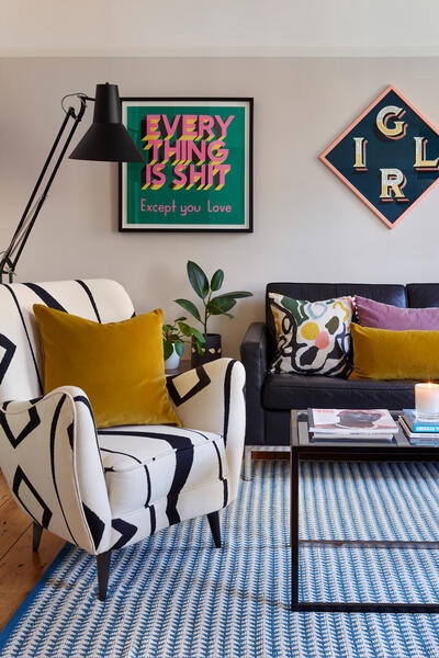 Armchair with geometric patterns sits beside a modern black lamp and leafy plant, in front of framed text: "EVERYTHING IS SHIT Except you Love". Background includes a sofa with pillows and wall art spelling "GIRL".