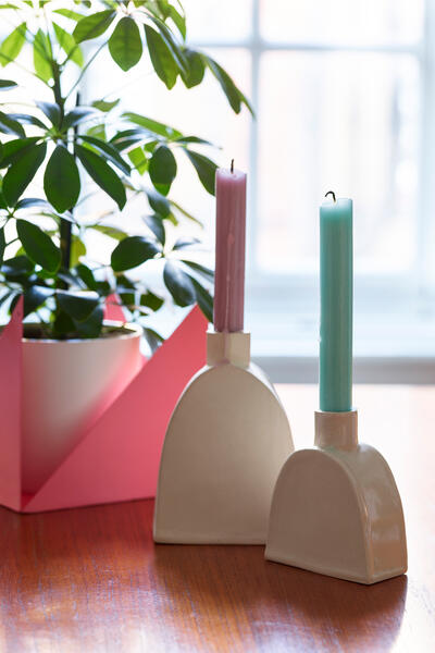 Pink and blue candles stand upright in abstract ceramic holders on a wooden table beside a green plant in a pink pot, with a bright window in the background.