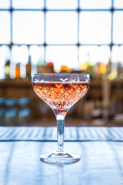 A crystal glass filled with a pink drink rests on a bar counter against a blurry background of a window and various bottles.