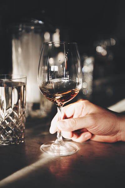 A hand holds a wine glass containing red wine on a dimly lit bar counter beside a crystal water glass, with blurry background lights.