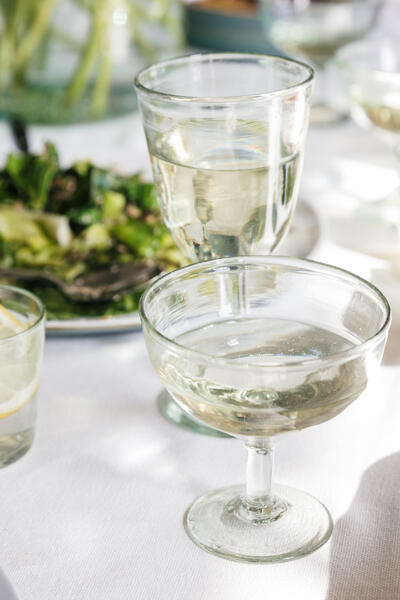 Two glasses of white wine and a water glass with a lemon slice on a white tablecloth, surrounded by a bowl of greens, utensils, and other glassware in a bright, sunlit dining setting.