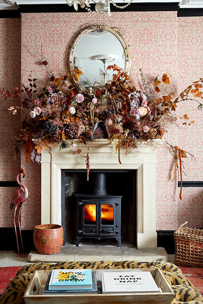 A wood-burning stove inside a fireplace with a floral arrangement above it, a mirror on the wall, and surrounding decorations including a flamingo statue, pot, books, and a zebra-patterned tray.