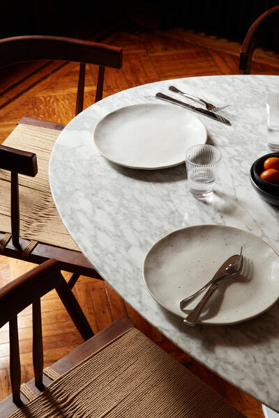 White marble dining table set with minimalist plates, utensils, and glasses; a black bowl of oranges; surrounded by wooden chairs with woven seats on a hardwood floor.