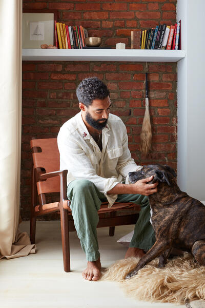 A barefoot man with dark hair sits in a chair petting a brindle dog, set in a cozy room with a brick wall, bookshelf, and large window with beige curtains.