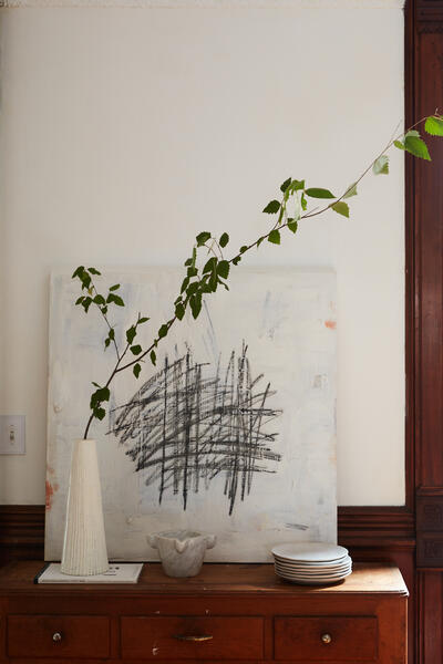 A long branch with green leaves in a tall, white vase sits on a wooden dresser. Behind it, there is a white abstract painting and a stack of white plates.