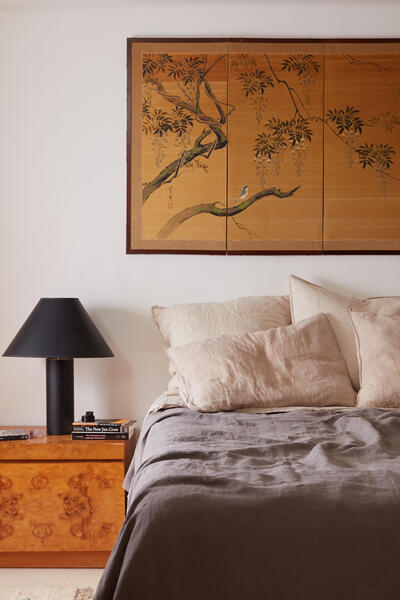 A bed with neutral pillows and a grey blanket is situated next to a wooden bedside table holding books and a black lamp, beneath a wall painting of a tree branch and bird. Text visible on a book cover includes: "The New Jim Crow."