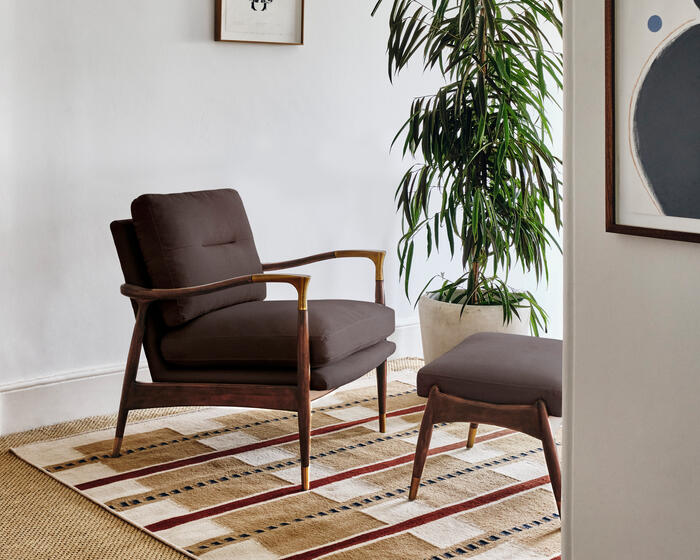 A brown armchair and matching ottoman sit on a patterned rug in a white-walled room, flanked by a potted plant and framed artwork.