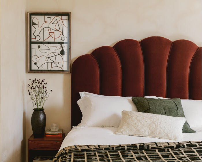 A modern bedroom features a bed with a maroon upholstered headboard, white and green pillows, and a patterned blanket. A wooden side table holds a dark vase with flowers and stacked books.