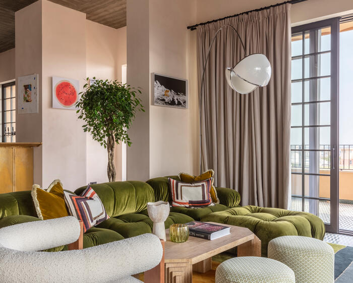 A modern living room with a green velvet sectional sofa, abstract-patterned pillows, and a wooden coffee table displaying books; a white armchair, green ottomans, floor lamp, and wall art add detail.