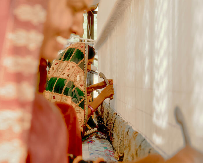 A woman in a patterned sari is working with a tool against a vertical woven fabric in an indoor setting with bright lighting. Another woman in the foreground appears out of focus.