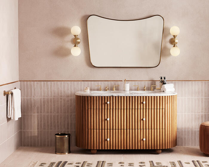 Wooden vanity with marble top, holding toiletries and rolled towels, against a tiled wall, below a large abstract-shaped mirror flanked by two globe lights in a modern bathroom.