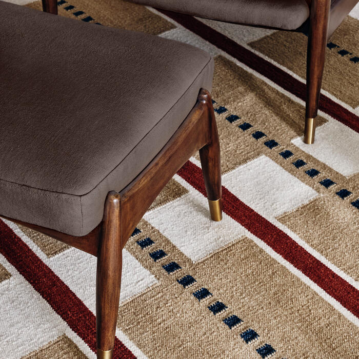 Wooden armchair and ottoman with brown cushions sit on a beige, red, and blue geometric-patterned rug in a living room setting.