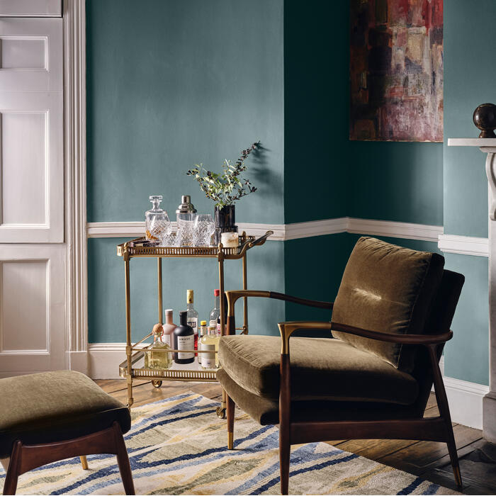 An olive-green armchair sits beside a stocked bar cart on a striped rug in a sophisticated room with teal walls, a white door, a marble fireplace, and a painting.