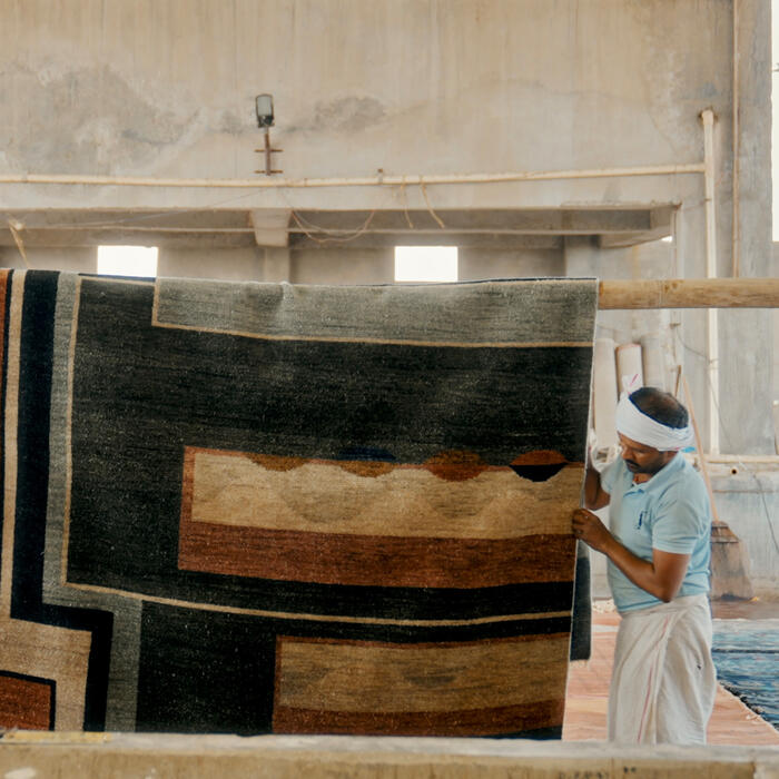 A person wearing a head wrap hangs a patterned carpet on a wooden frame inside an open, industrial warehouse with concrete walls and pigeons perched near the ceiling.