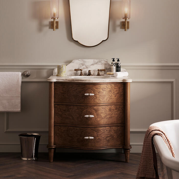 A wooden vanity with a marble countertop supports a mirror and two wall-mounted light fixtures, set in an elegant bathroom featuring a towel holder, wastebasket, and partial view of a bathtub.