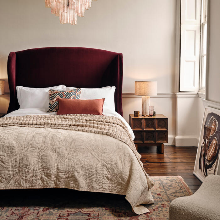 A plush bed with a burgundy headboard sits centered under a cascading crystal chandelier in a cozy bedroom. Flanked by wooden nightstands, one with a lamp, the room features a patterned rug and soft, neutral decor.