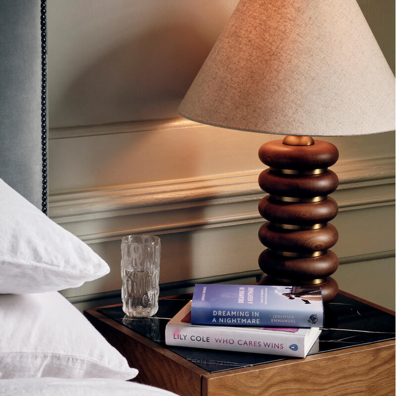 A wooden bedside table holds a modern lamp, glass of water, and two books titled "Dreaming in a Nightmare" by Jeremiah Emmanuel and "Who Cares Wins" by Lily Cole, next to a bed.