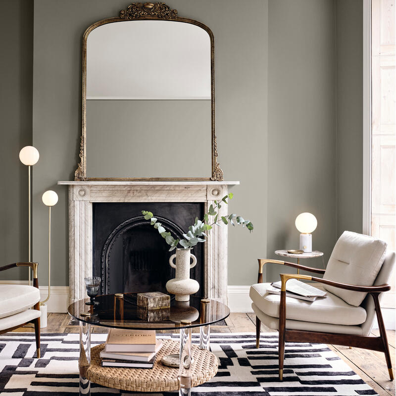 Two armchairs face each other beside a fireplace with a large mirror; round coffee table with books and vase in a cozy, elegant living room with geometric rug and warm lighting.