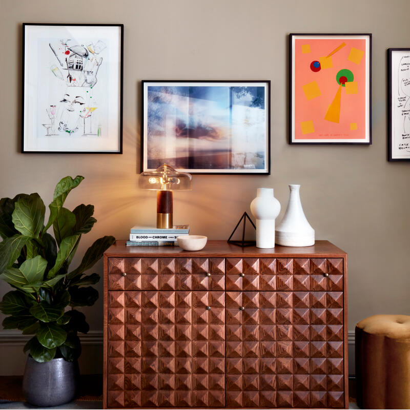 A wooden cabinet with geometric patterns holds white vases and books beneath a lamp in a room with wall art, a potted plant in a gray pot, and decorative moldings.