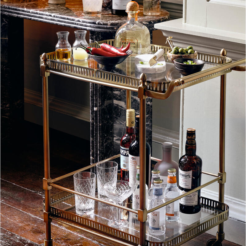 A brass bar cart holds various bottles, glassware, and food items, situated in a stylish room characterized by a marble counter and wooden floor.