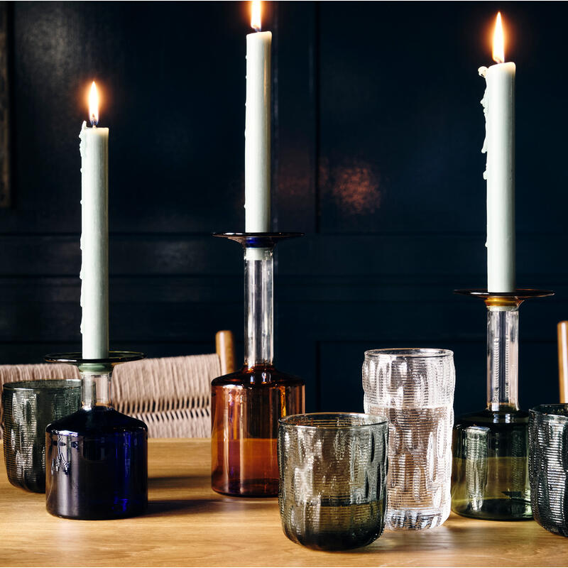 Lit candles stand upright in glass bottles on a wooden table, surrounded by empty drinking glasses, in a dimly lit room with dark walls and wooden chairs.