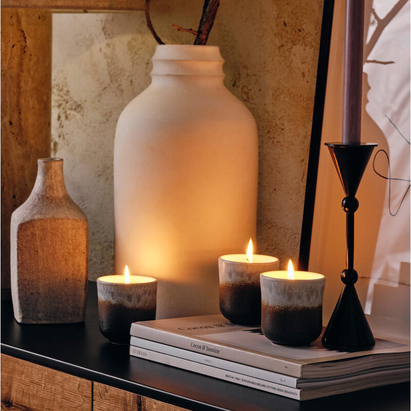 Lit candles rest on magazine stacks atop a dark wooden surface near large vases and a framed artwork, creating a rustic, cozy atmosphere.

Text on magazines: "Cocoa & Jasmine, LUMFEAS, CARRIERE ..." (partial text).