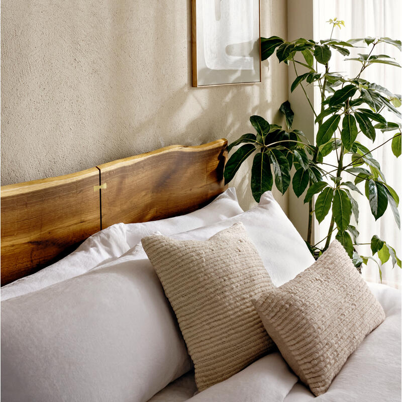 Bed with white linens and beige pillows, situated against a textured beige wall, adorned with abstract framed art, beside a leafy plant near a window with sheer curtains.