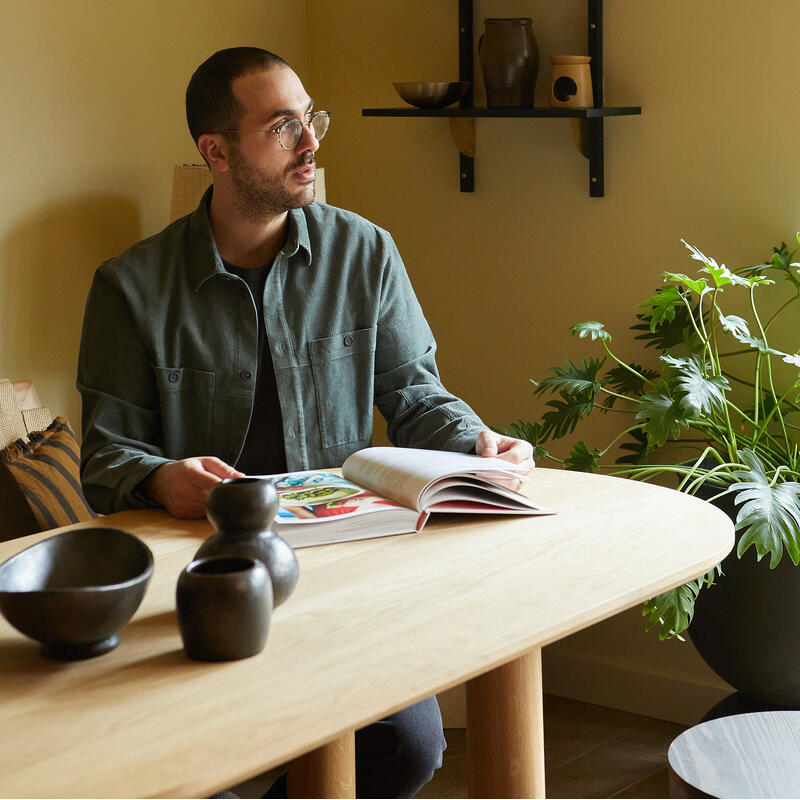 A man sits at a wooden table reading a book in a cozy room with beige walls, decorative shelves, and several potted plants.